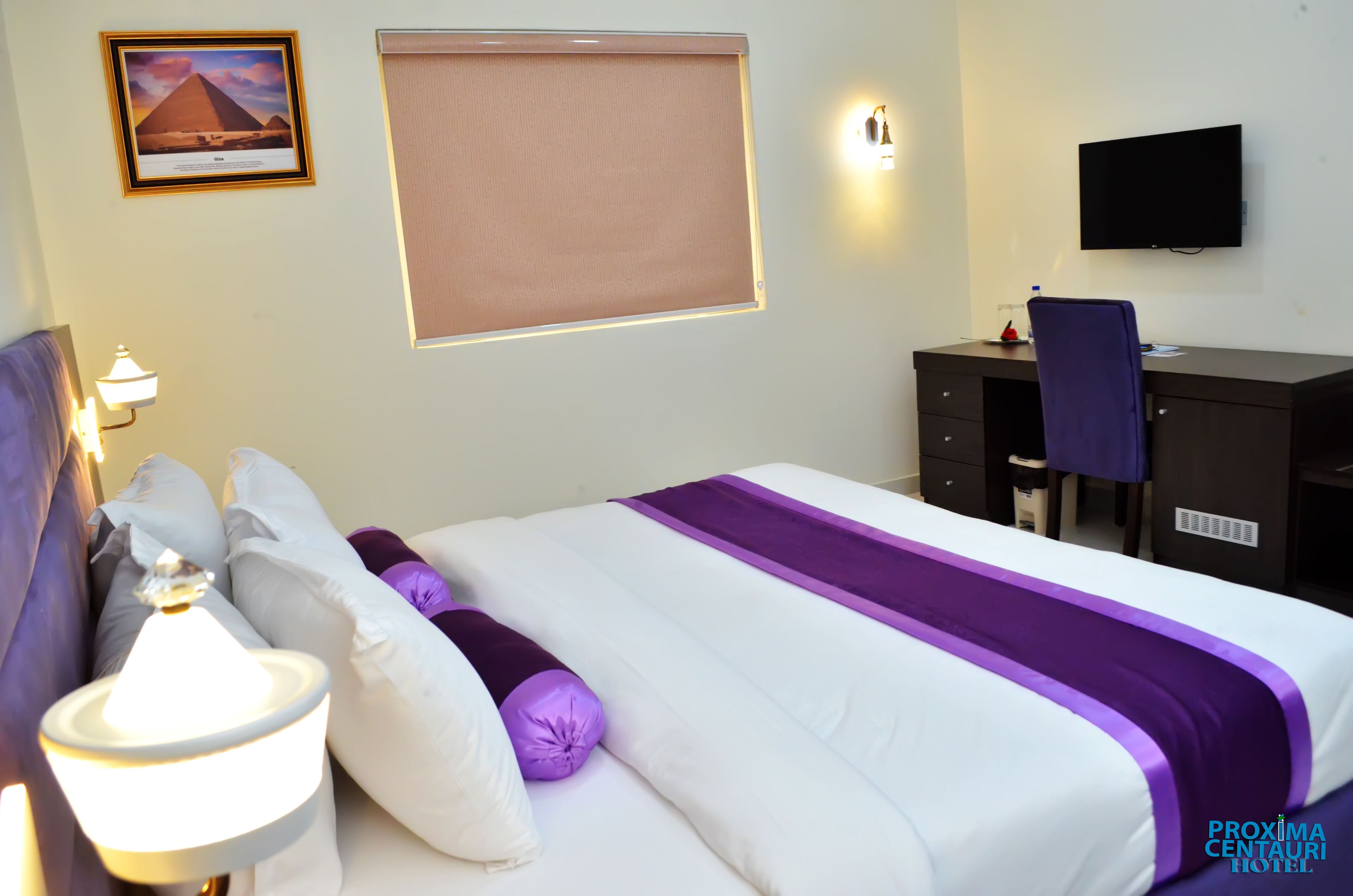 A hotel room with a bed, a desk, a chair, a TV, a lamp, and a picture of a pyramid.