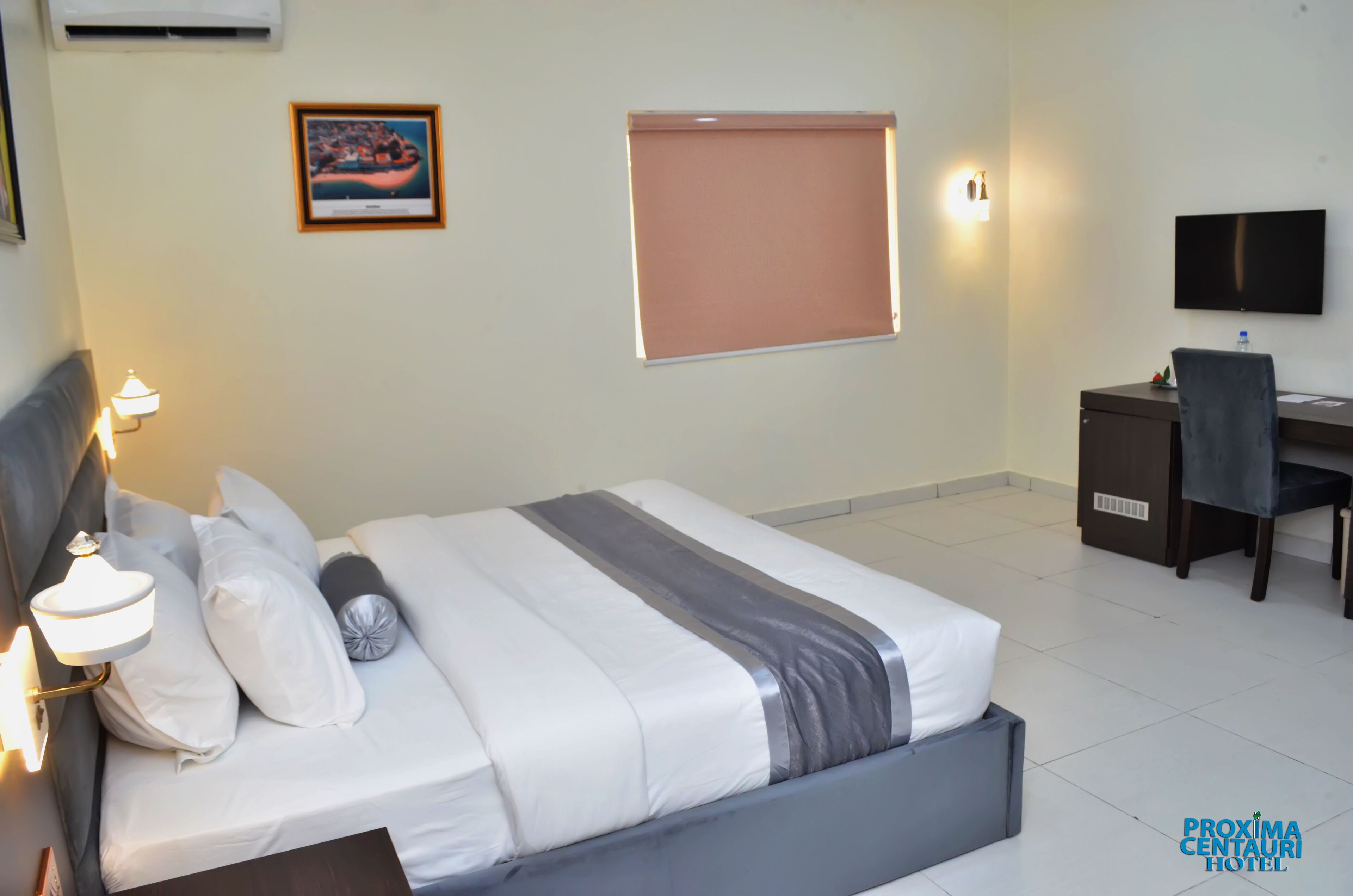 A hotel room with a bed, bedside lamp, desk, chair, TV, and a framed picture on the wall.