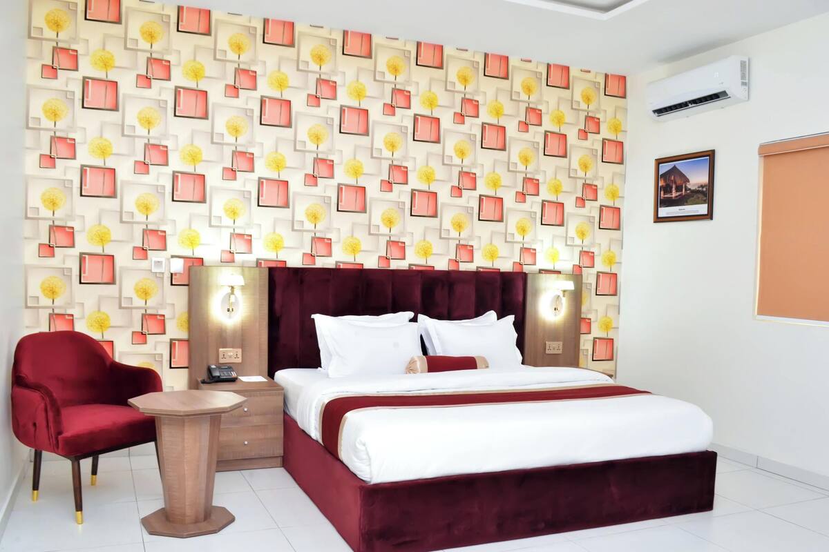 Habitación de hotel con pared estampada, una silla roja, una mesita de noche, una cama con ropa blanca y un cuadro en la pared.