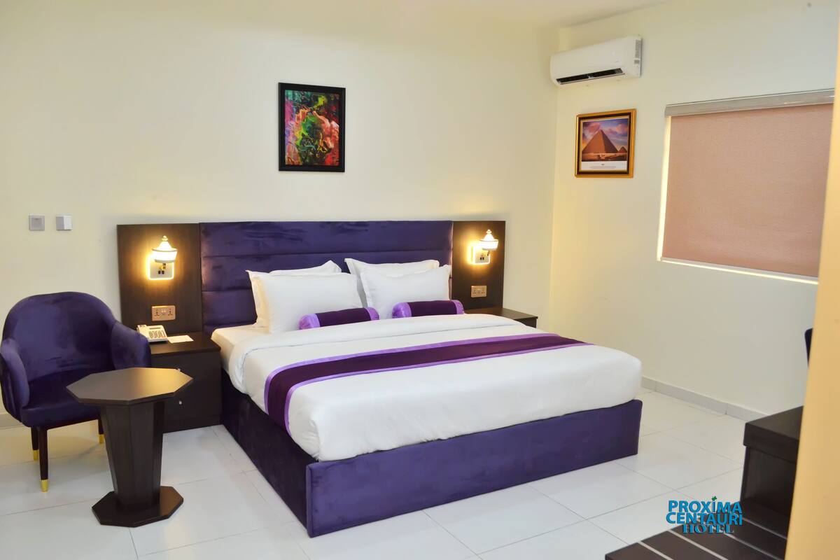 Una habitación de hotel con una cama grande, dos mesitas de noche con lámparas, una silla y obras de arte en las paredes.