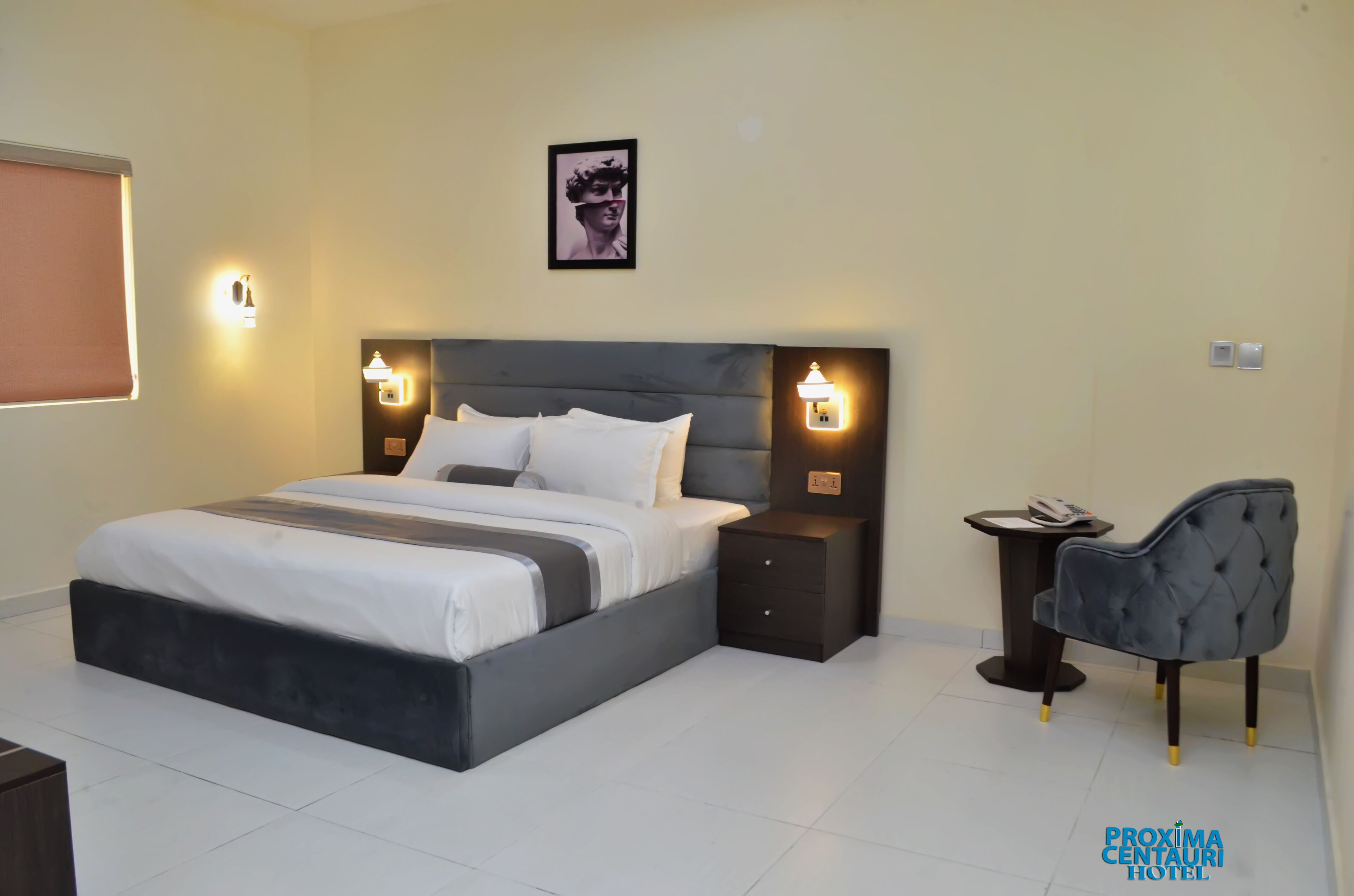 A modern hotel room with a large bed, bedside tables, a chair, and a framed picture on the wall.