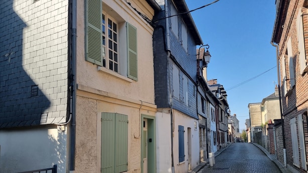 A narrow cobblestone street lined with historic buildings featuring traditional European architecture, including shuttered windows and brick facades.