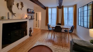 A modern living room with a fireplace, wooden flooring, a dining area with a table and chairs, and large windows with curtains.