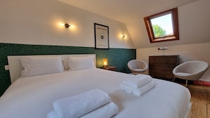 A neatly made bed with white linens and towels, a framed poster on the wall, a small table with a lamp, and a window with a view of trees.