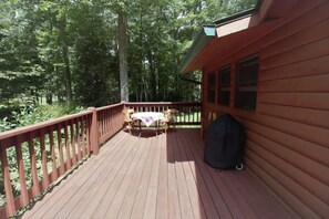 A wooden deck with a table and chairs, surrounded by a wooden railing, set in a forested area. - Sunny and Cozy Creekside Cabin (Banner Elk)