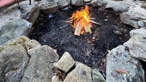 Ein Steinkamin mit Holzscheiten, umgeben von Steinen, in einer bewaldeten Gegend mit einer Hütte im Hintergrund.