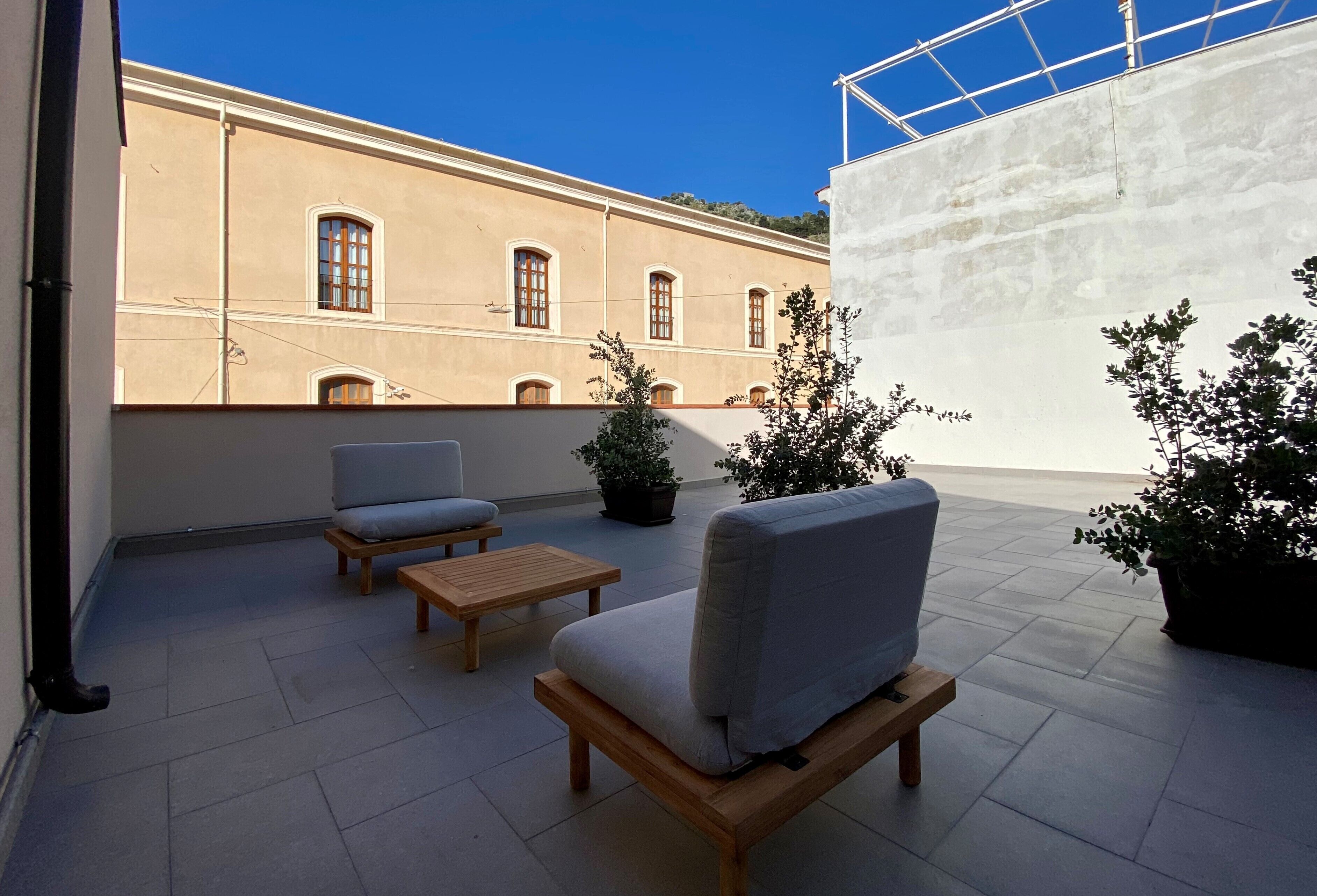 Una terraza con dos sofás y una mesita, un edificio color beige con ventanales arqueados y plantas en macetas.