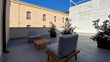 Una terraza con dos sofás y una mesita, un edificio color beige con ventanales arqueados y plantas en macetas.