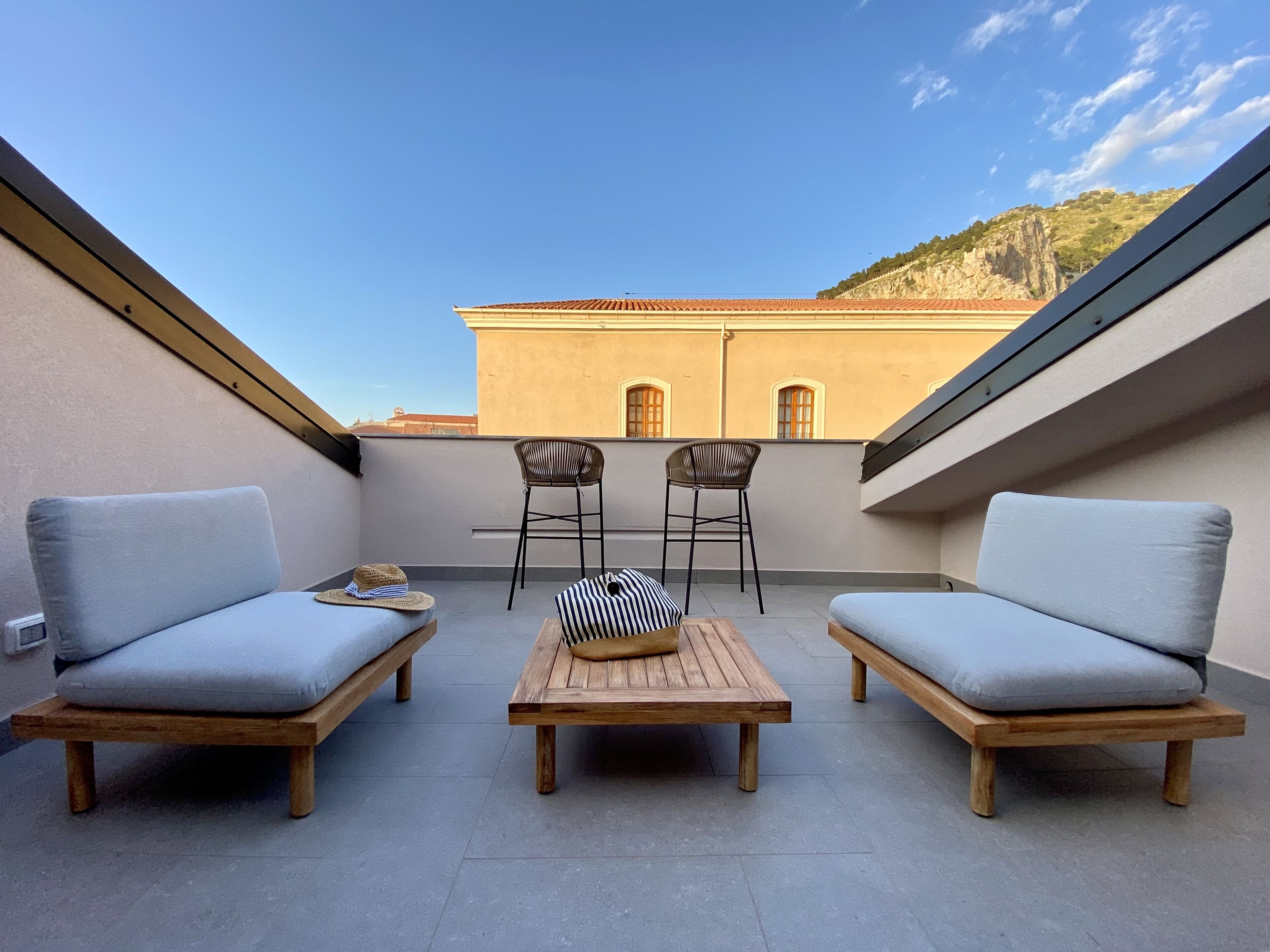 A rooftop terrace with two sofas and two chairs, a wooden table, and a view of a building and hills in the background.