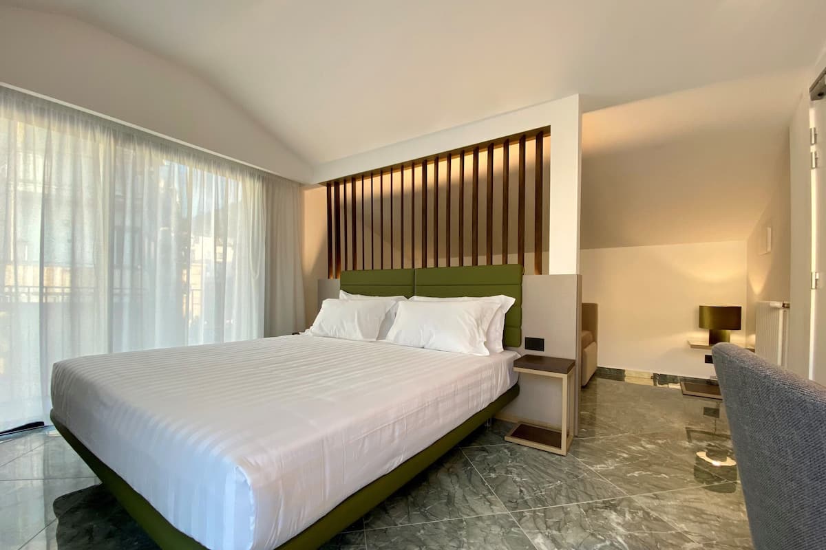 A modern hotel room with a large bed, a nightstand, a chair, and a view of the city through large windows.
