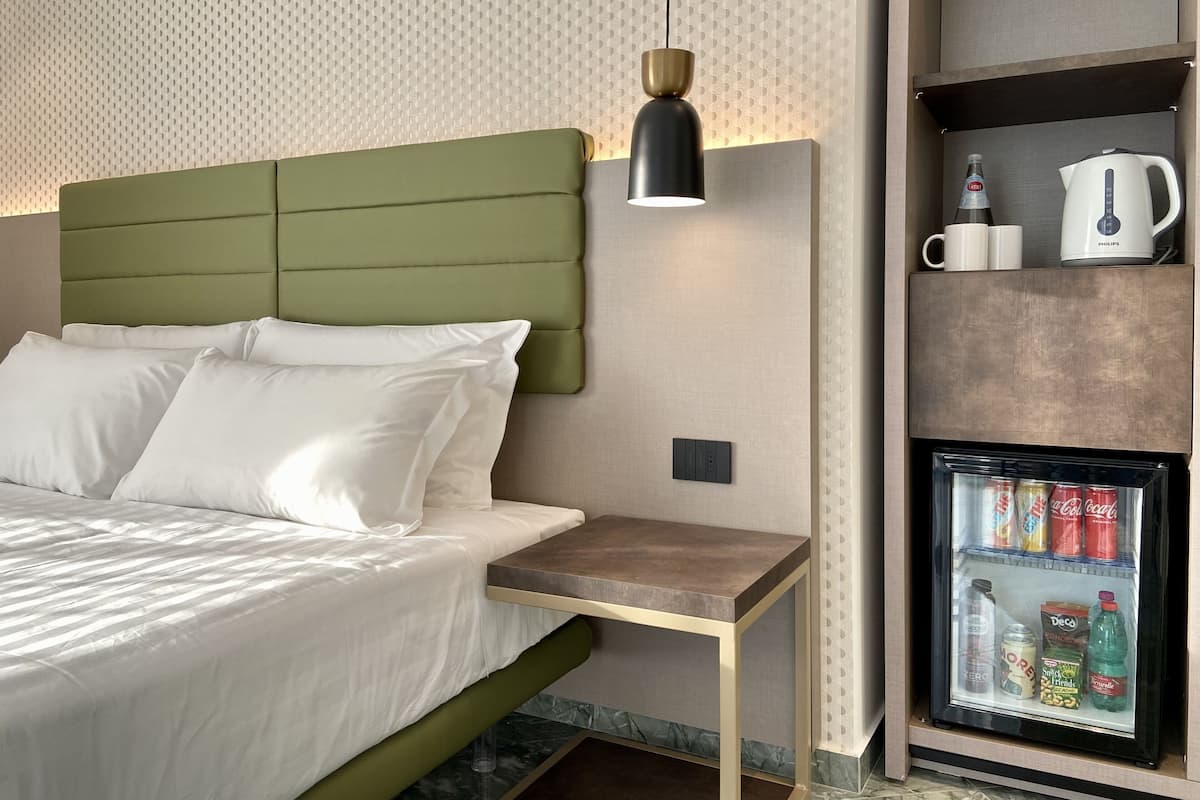 A modern hotel room with a green upholstered headboard, a bedside table, a lamp, and a minibar with beverages and snacks.