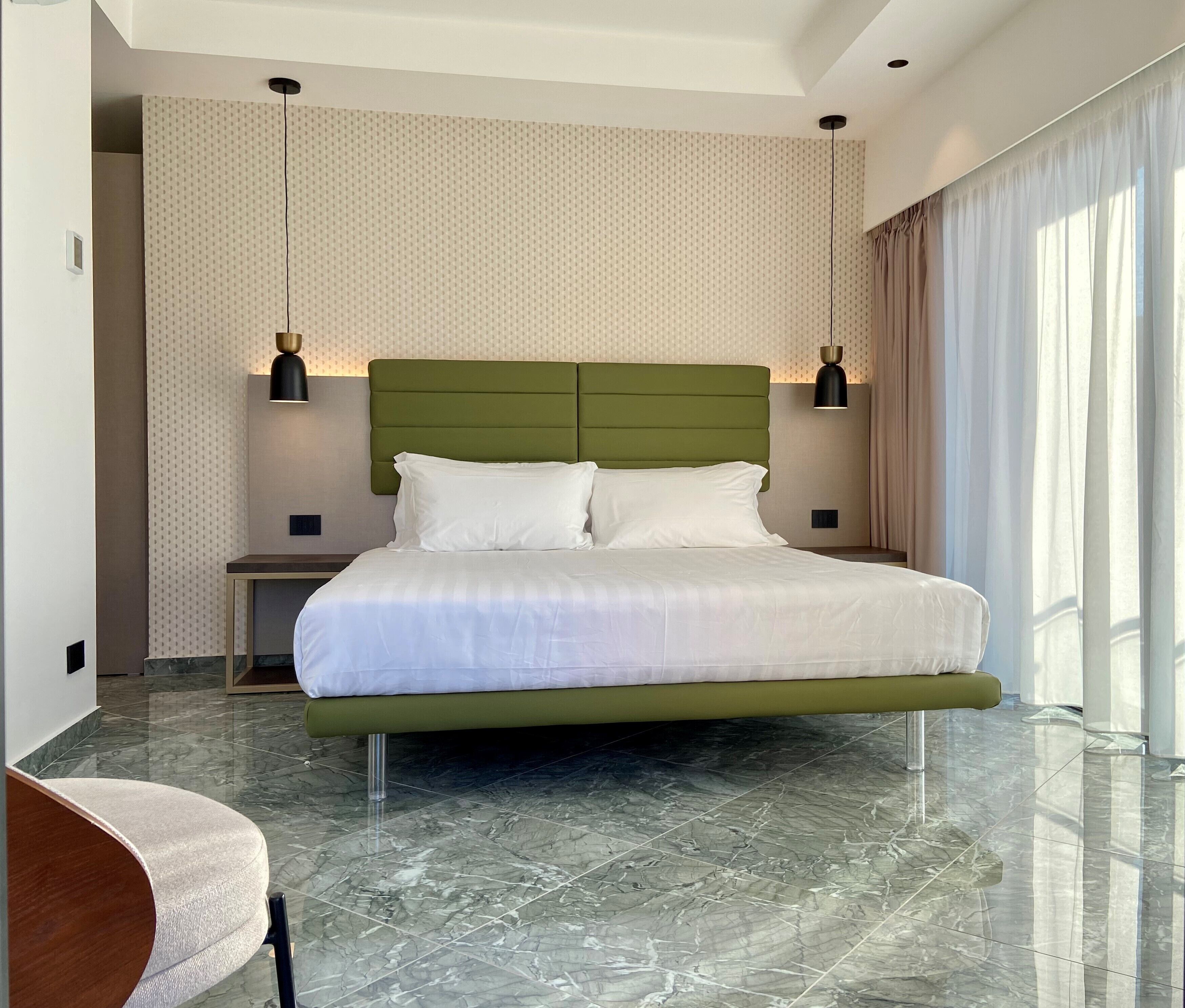 A modern hotel room with a green upholstered headboard, a large bed with white linens, two bedside tables with lamps, and a marble floor.