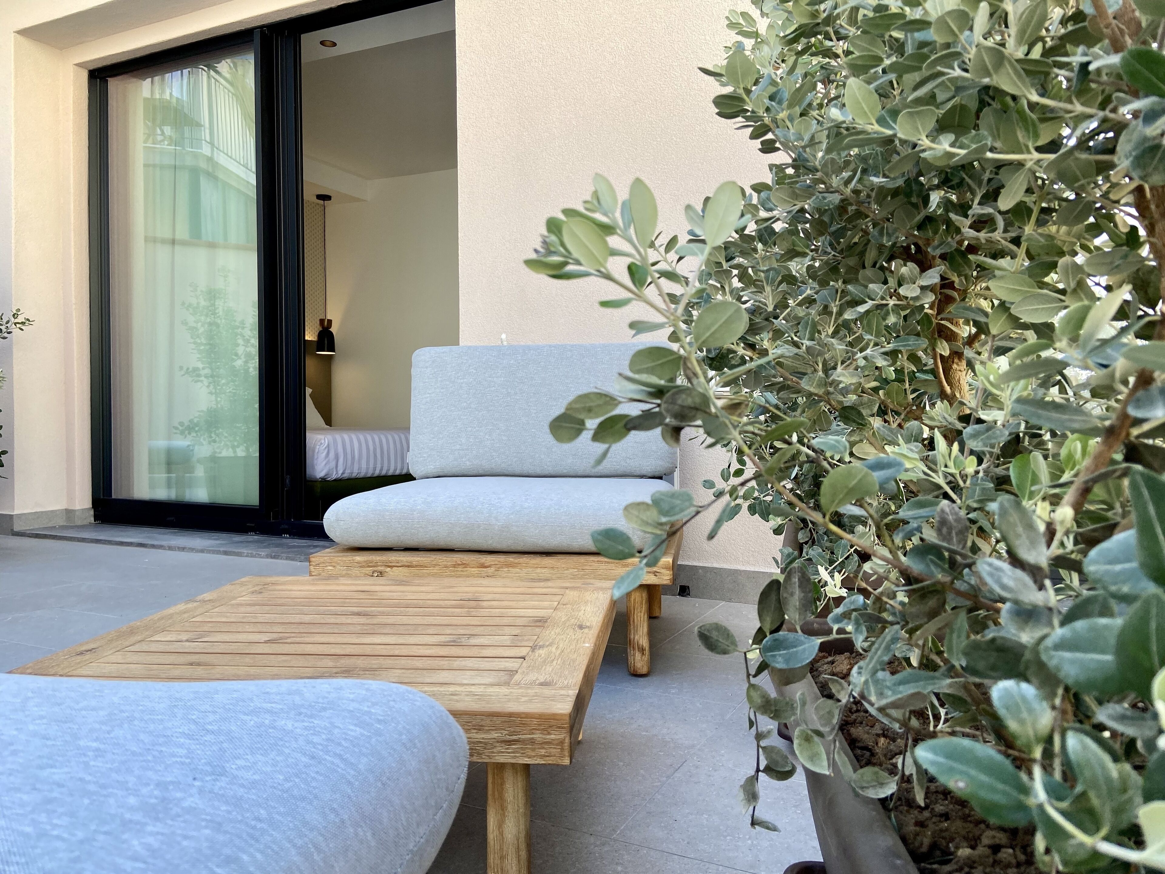 A modern outdoor seating area with a wooden table and two cushioned chairs, a potted plant, and a sliding glass door leading to a bedroom.