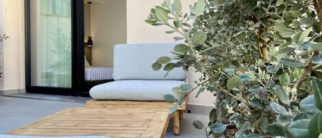 Un espace extérieur moderne aménagé avec une table en bois et deux fauteuils rembourrés, un pot de fleurs et une porte-fenêtre coulissante donnant sur une chambre.