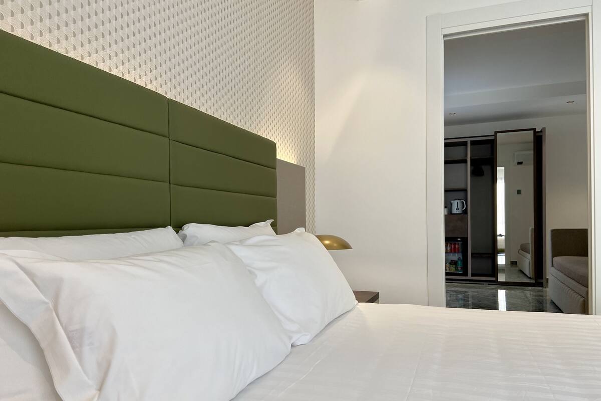 A neatly made bed with white linens, a green upholstered headboard, and a bedside lamp.