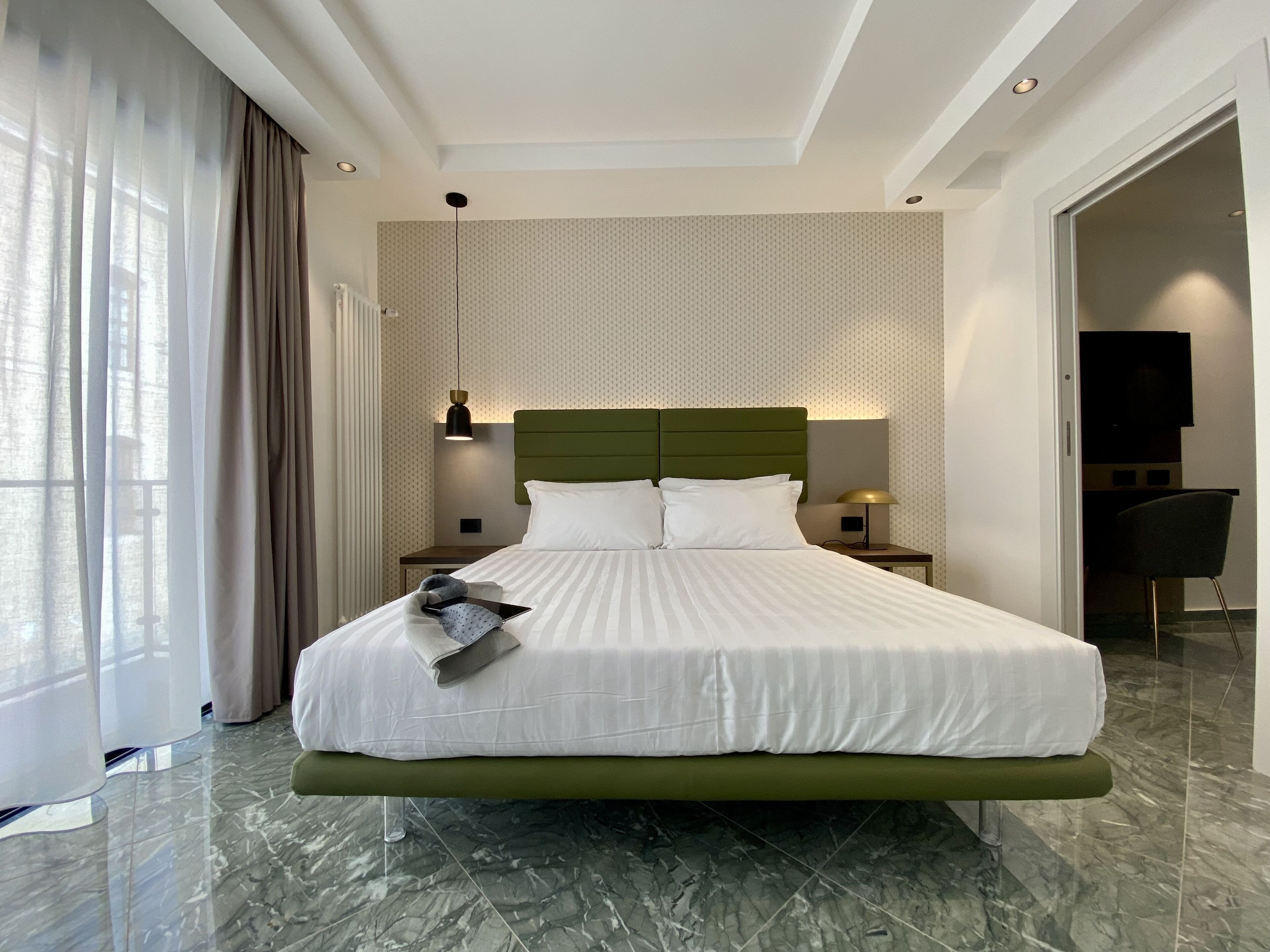 A modern hotel room with a large bed, a green headboard, a nightstand with a lamp, and a view of the city through large windows.