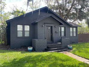 A single-story house with a dark gray brick exterior, a front door with a window above it, and a small porch with steps leading up to the door. There are two planters on the front steps. - Mid-Century Home:  Backyard, Close to Downtown (Jacksonville)