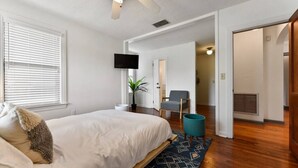 Ein Schlafzimmer mit einem Bett, einem Fernseher, einem Sessel, einer Pflanze und einem Flur, der in einen anderen Raum führt.