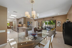 A modern kitchen with a central island, a dining area with a glass table, and a living room with a sofa and chairs.
