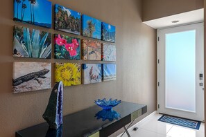 A hallway with a series of framed artworks on the wall, a glass table with a blue vase, and a door with a glass panel. - Palm Springs, Greenhouse East! 2 Bdrs/2 Baths/ 25 Yard pool/Amazing location! (Palm Springs)