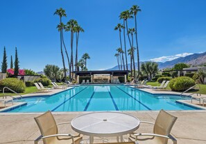 A swimming pool with lounge chairs and a table in the foreground, surrounded by palm trees and a mountain backdrop.