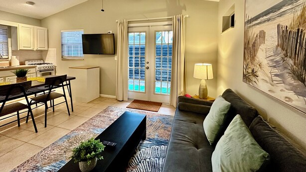 A modern living room with a sofa, a coffee table, and a TV. There is a kitchen area with cabinets and a stove. A painting is on the wall, and a window with curtains is visible. - CHARMING INDEPENDENT GUEST HOUSE (West Palm Beach)