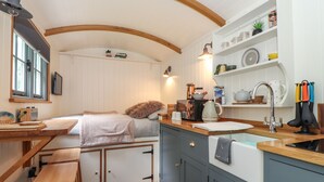 Een kleine keuken met houten kastjes, een spoelbak en een aanrecht. Boven de spoelbak is een raam en er is een plank met keukengerei.