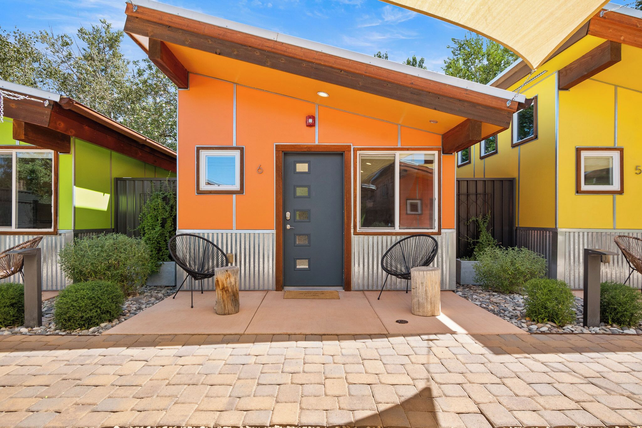 A building with a modern design, featuring a prominent orange facade, a dark blue door, and a small porch area with two chairs.