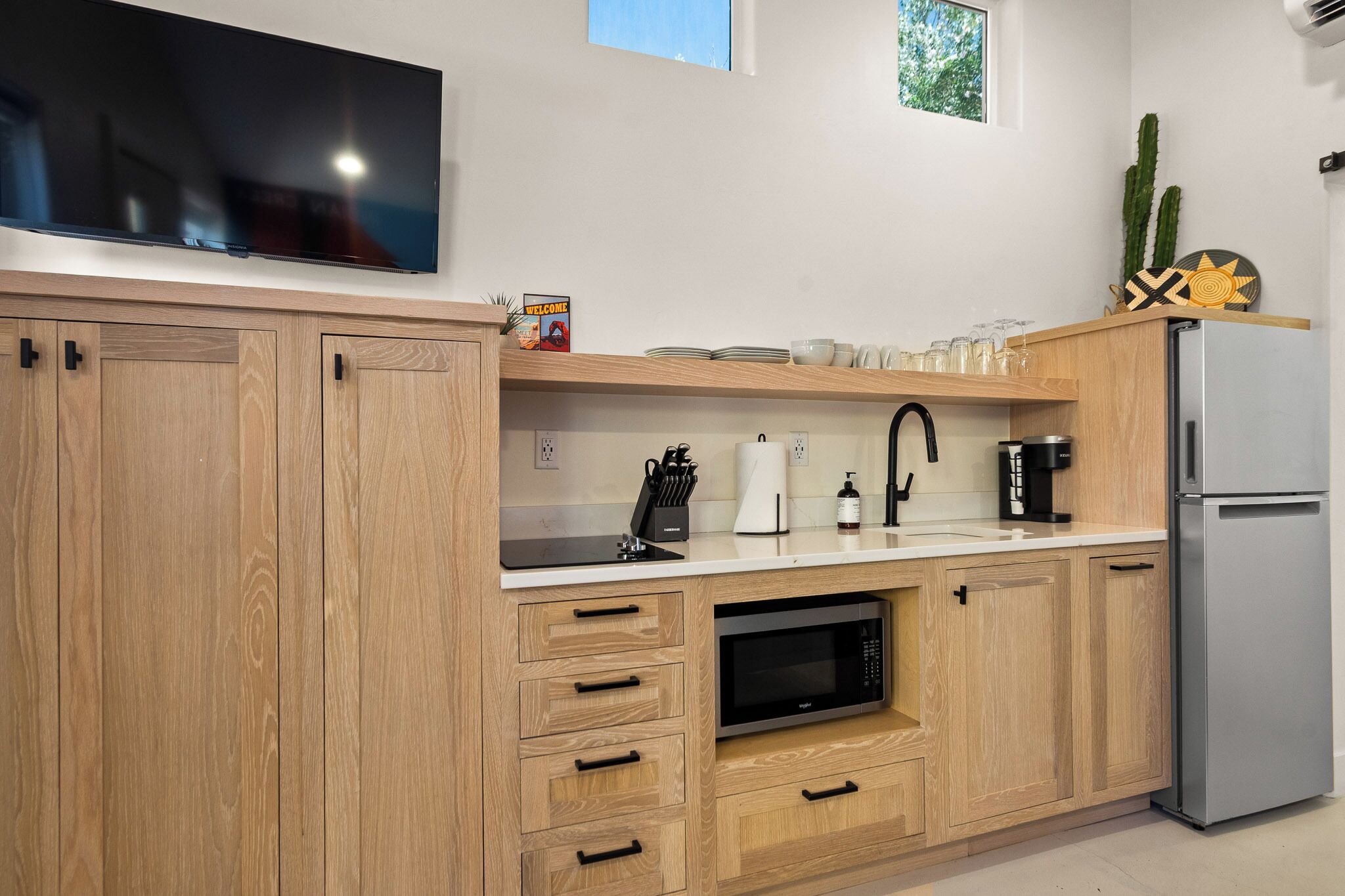 A modern kitchen with wooden cabinets, a built-in microwave, a stainless steel refrigerator, and a flat-screen TV mounted above the counter.
