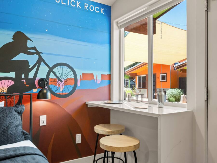 A room with a mural of a cyclist, a bed, a window with a view of buildings, and two stools.