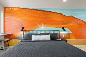 Un dormitorio con un mural de atardecer, una cama con ropa de cama gris y un escritorio con una silla.
