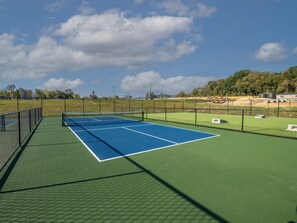 An outdoor tennis court with a green surface and a blue playing area, surrounded by a fence.