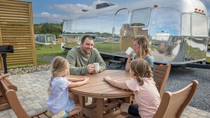 A family of four seated at a round wooden table outdoors with a trailer in the background.