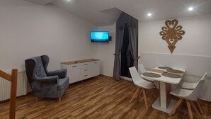 A modern living room with a wooden floor, a grey armchair, a white dining table with four chairs, a flat-screen TV mounted on the wall, and a decorative wall piece. - Lovely Mountain Apartment Zakopane 14km (Suchá Hora)