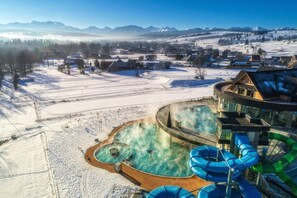 Un complejo turístico con zona de aguas termales, tobogán de agua y vistas a montañas nevadas.