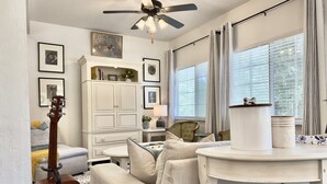Un salon avec un ventilateur de plafond, un canapé, une guitare, un meuble contenant des objets décoratifs et une fenêtre avec des rideaux.