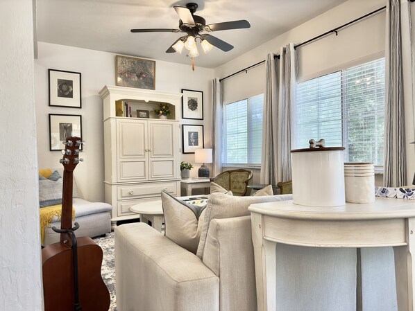 A living room with a ceiling fan, a couch, a guitar, a cabinet with decorative items, and a window with curtains.