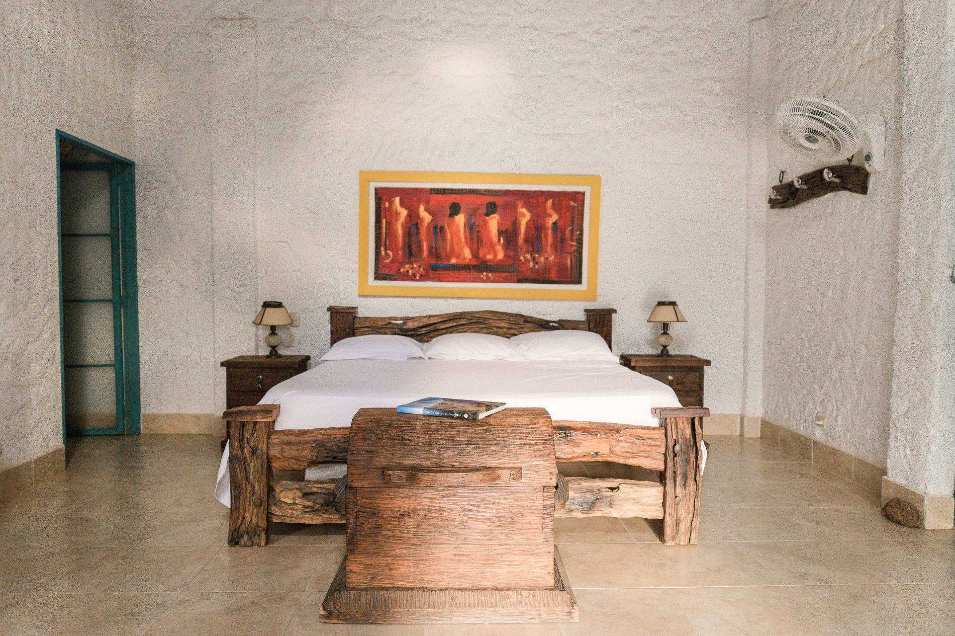 Un dormitorio con estructura de cama de madera, colchón blanco, dos mesitas de noche con lámparas, un ventilador de pared y un cuadro sobre la cama.