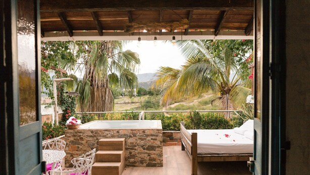 An outdoor patio with a stone wall, a wooden bench, a white bed, and a white chair with pink cushions. - HOTEL KALMA  (Santa Fe de Antioquia)