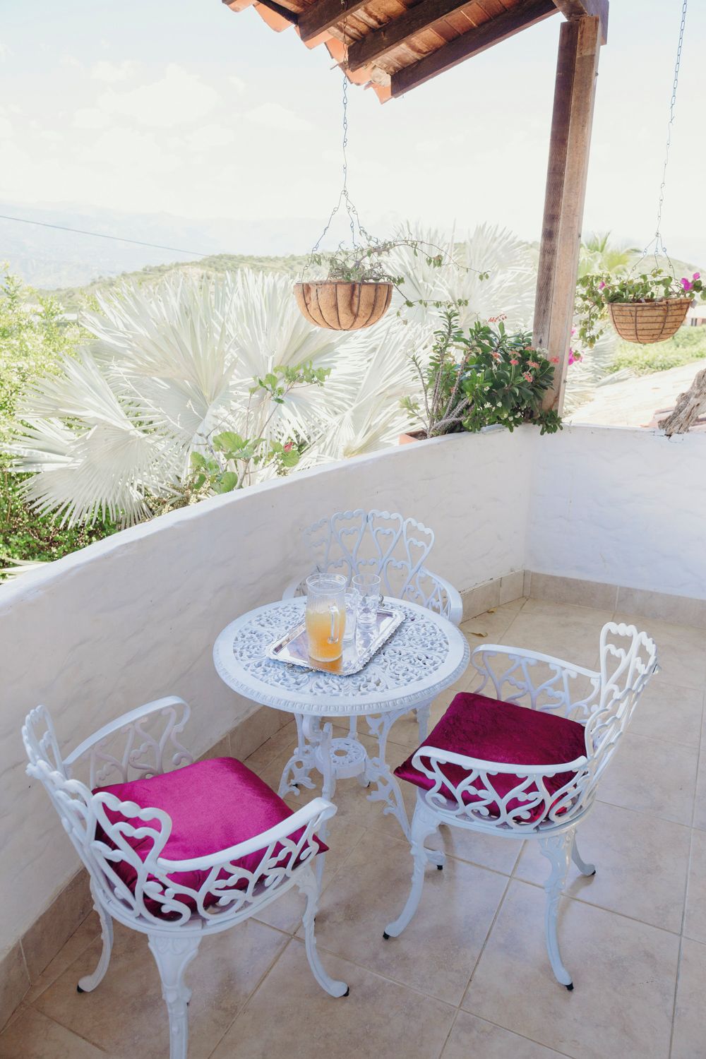 A patio with a round table and two chairs, a hanging planter, and a view of greenery.