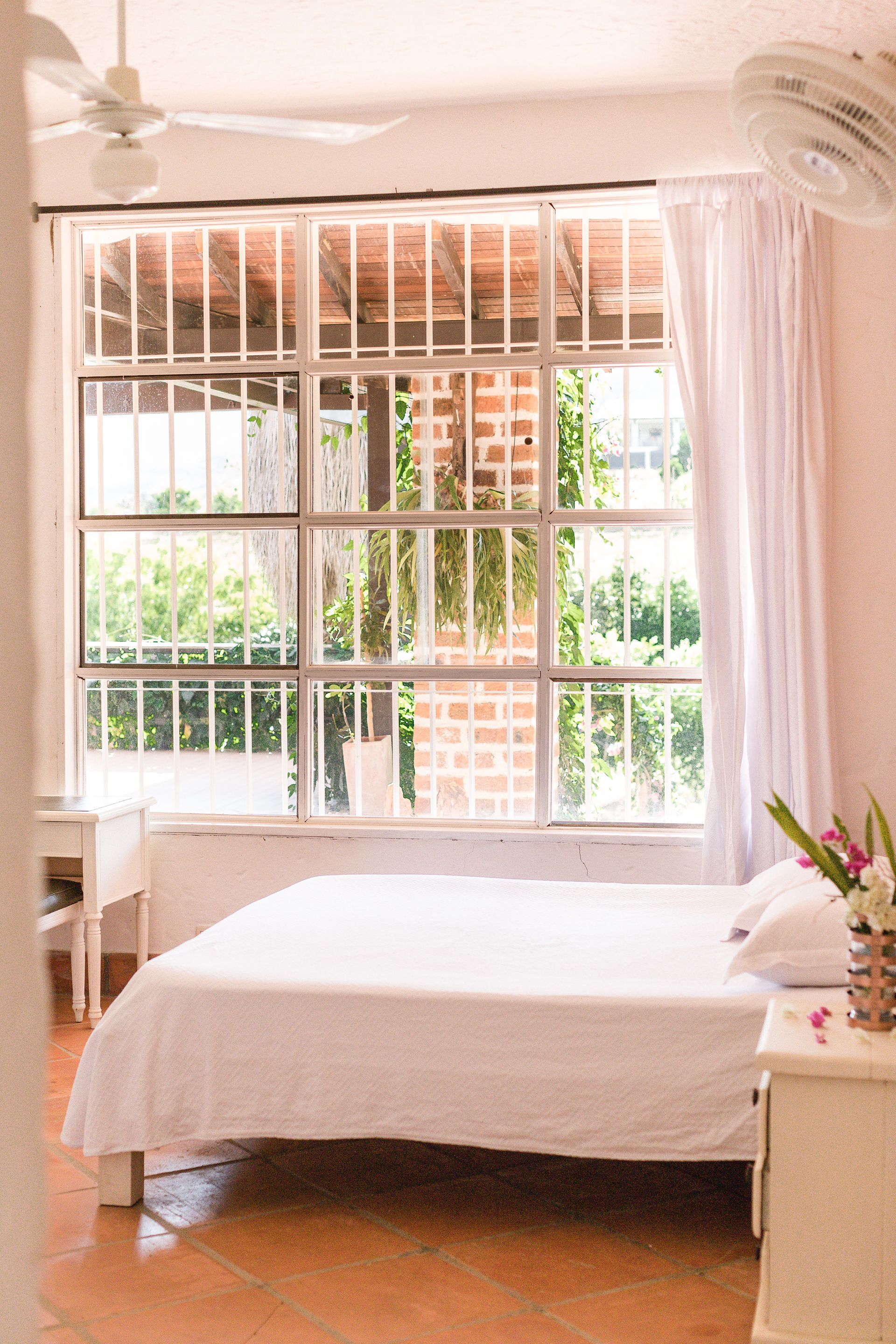 A bedroom with a bed, a nightstand, a window with a view of greenery, and a ceiling fan.