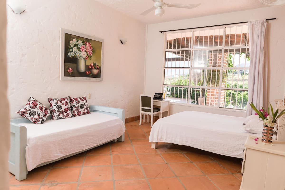 A room with two beds, a desk, a chair, a ceiling fan, and a window with bars.