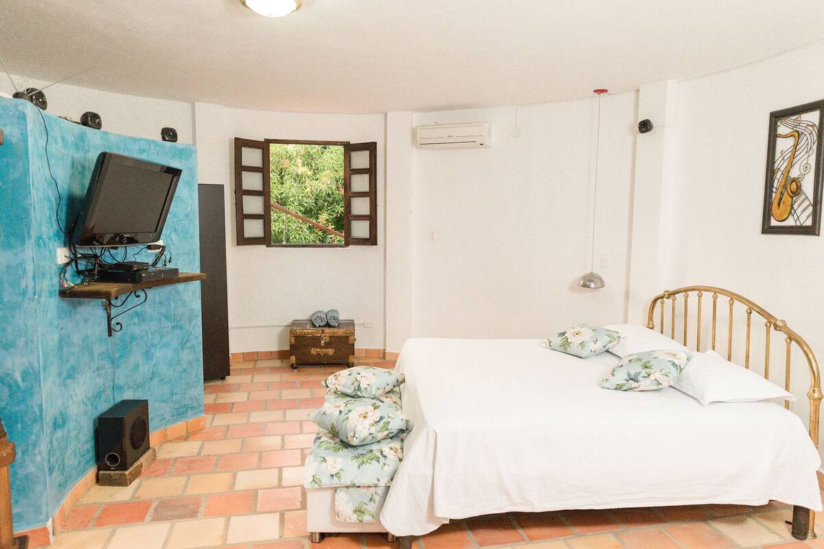 A bedroom with a bed, a television on a stand, a window with shutters, a chest of drawers, and a wall-mounted air conditioner.