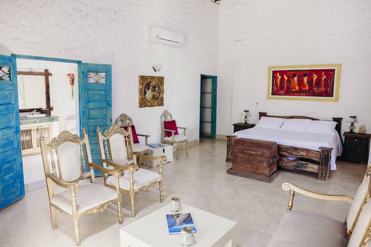 A room with a bed, two chairs, a table, a picture frame, and a door.
