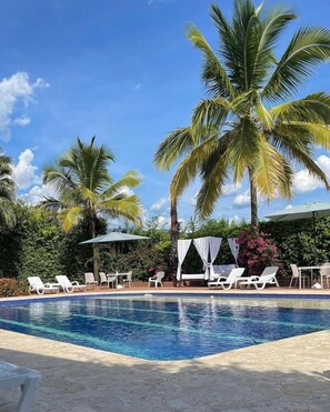 Un área de piscina con sillas reclinables blancas y un espacio de estar techado, rodeado de palmeras y follaje.