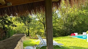 A covered outdoor seating area with a concrete bench and a wheelbarrow, overlooking a pool and lush greenery.