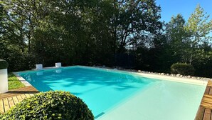 A rectangular swimming pool with a wooden deck surrounding it, set in a landscaped garden with trees and a neatly trimmed hedge.