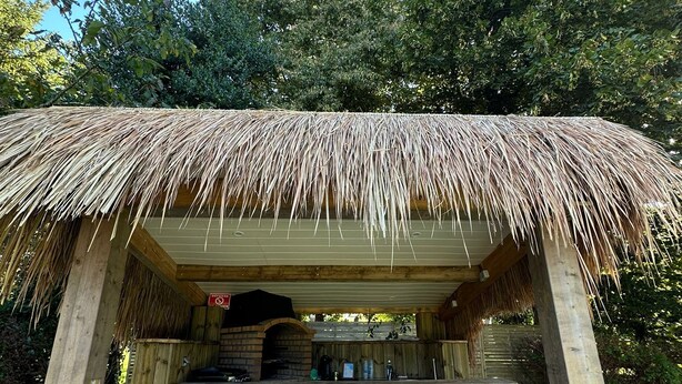 A rustic outdoor bar with a thatched roof, wooden stools, and a brick oven.