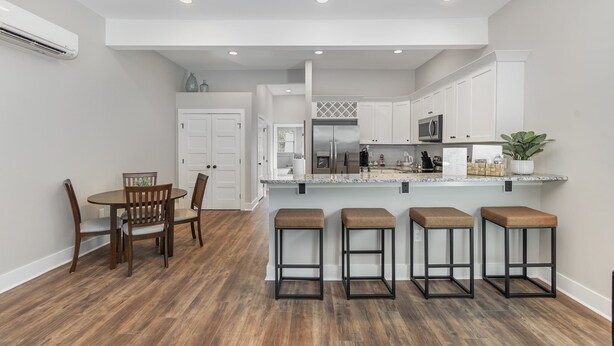A modern kitchen with white cabinets, a central island with stools, and a dining area with a wooden table and chairs. - Modern Condo in Heart of Downtown Fuquay (Fuquay-Varina)
