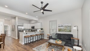 A modern living room with a gray sofa, a coffee table, and a dining area with wooden chairs and a bar stool. - Modern Condo in Heart of Downtown Fuquay (Fuquay-Varina)
